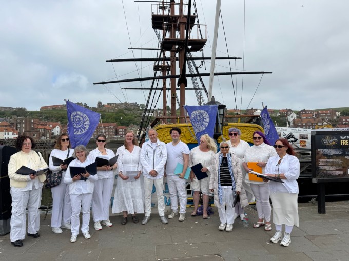 The Whitby Community Choir performing a cappella at Dock End during the Fish & Ships Festival, dressed in white attire, with a historic ship and colorful flags in the background.